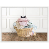 Black cat peeking over a pile of pastel-colored laundry in a woven basket on a wooden floor with paw prints leading away toward a white paneled wall.