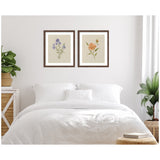 Two framed botanical prints above a white bed, featuring purple and orange flowers on beige backgrounds in simple wooden frames, flanked by green plants.