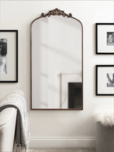 Tall rectangular wall mirror with an arched top and ornate carved wood frame hanging between two black-framed photos above a light-colored sofa and chair.