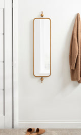Tall rectangular mirror with rounded corners and brass metal frame, mounted vertically on white wall, with beige robe hanging nearby and brown slippers below.