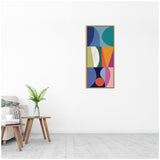 Framed vertical abstract canvas with geometric shapes in blue, green, orange, purple, and pink, hung on a white wall above a wooden side table and beige sofa.