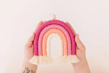 Hands holding a handcrafted rainbow decoration made of yarn in shades of pink, peach, and cream, with fringed ends and a small wooden bead on top.