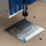 Close-up of a PROVerXL 4030 CNC router engraving 'Geomillkstl PROVerXL 4030' on a reflective metal plate, shown under a blue spindle on a wooden base.