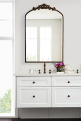 Rectangular wall mirror with an ornate dark bronze frame and decorative crest above, hung above a white bathroom vanity with brown knobs and floral arrangement.