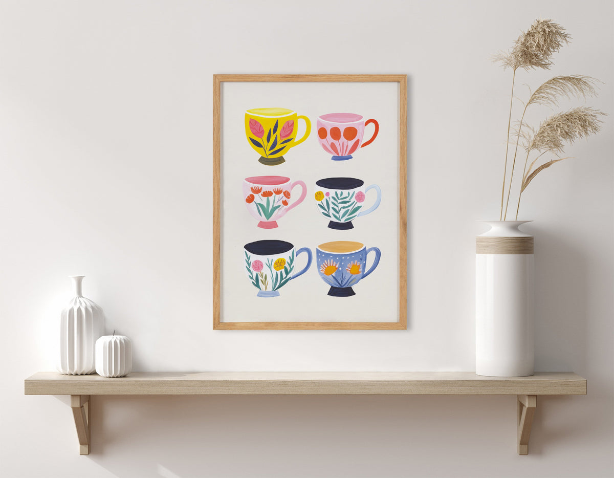 Framed print of six colorful coffee cups with floral patterns arranged in two columns, displayed above a wooden shelf with white vases and dried pampas grass.