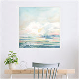 Soft pastel watercolor painting of a cloudy sky over a serene landscape with green, blue, and pink hues, framed and hung above a light wood desk.
