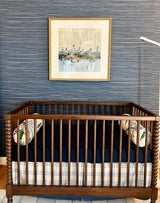 Dark wood crib with vertical spindles and patterned bolster pillows set against textured blue wallpaper, framed duck-themed fine art print above the crib, and a white adjustable lamp to the side.