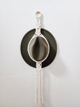 Olive green felt hat hanging on a white macrame hanger with braided patterns and long hanging fringe against a white wall.