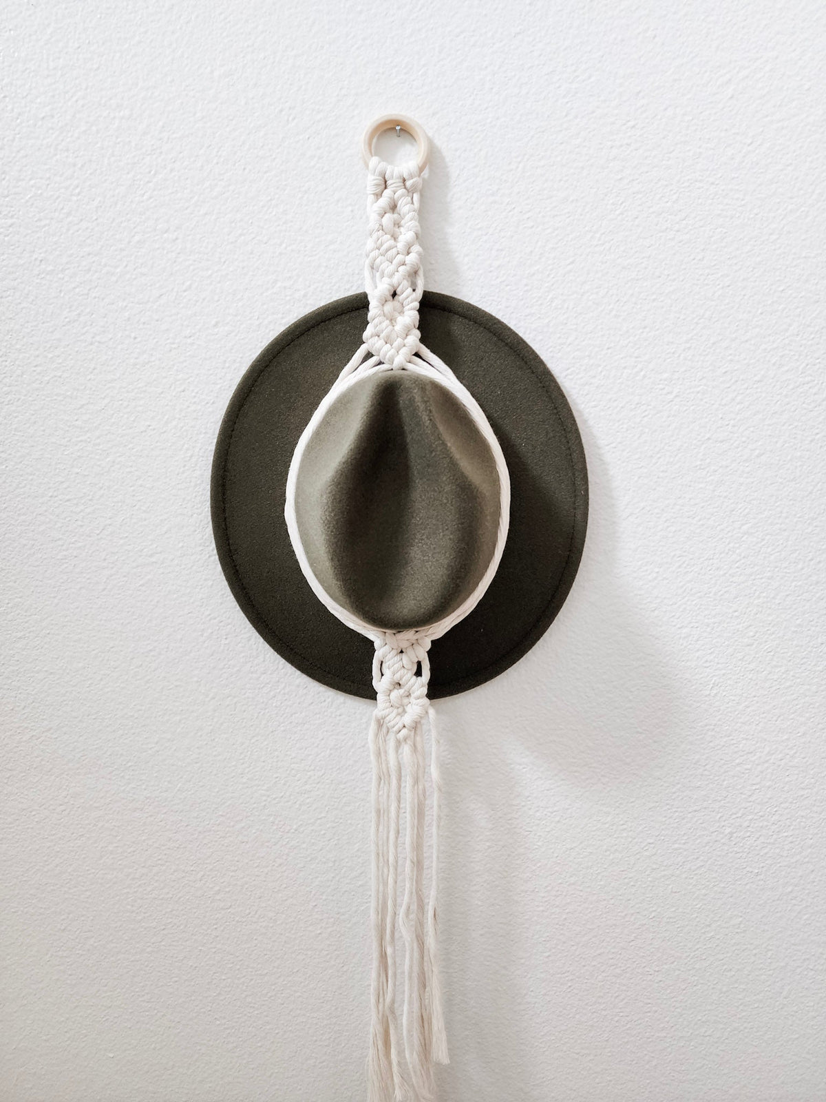 Olive green felt hat hanging on a white macrame hanger with braided patterns and long hanging fringe against a white wall.