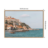 Coastal village with pastel-colored buildings on a rocky hillside beside teal ocean waters under a pale blue sky with a single cloud, framed with a light wooden border.