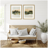 Two framed abstract landscape prints with soft beige and green tones hanging side by side above a wooden bench with white cushion and black-white plaid pillows, flanked by a large potted plant.