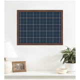 Rectangular framed fabric pinboard with navy blue and light beige plaid pattern mounted on a white wall above a white shelf with a lamp, photo frame, and a potted plant.