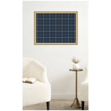 Navy blue fabric pinboard with light wood frame and thin tan grid lines, hanging on white wall above two curved cream chairs and a small brass side table with a white vase.