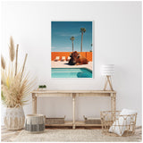 Framed photograph of a large bison lying beside a bright blue pool with two tall palm trees against a clear sky and an orange wall in the background, displayed above a wooden console table.