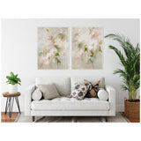 Two soft pastel floral canvas paintings with muted pink, green, and beige hues framed in white, hung above a white sofa with patterned and solid pillows in a minimalist living room.