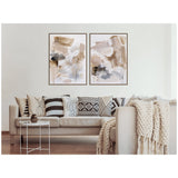 Two framed abstract canvas artworks in beige, tan, black, and white hang above a beige sofa with patterned black and white pillows and a chunky knit throw in a minimalist living room.