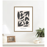 Framed abstract art print titled 'Les Fleurs' with black organic shapes on a beige background hanging above a white surface with a lamp, photo frame, and potted plant.
