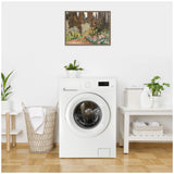 Framed impressionist garden painting with green, brown, and pink hues hanging above a white front-load washing machine in a minimalist laundry room with plants and a basket.