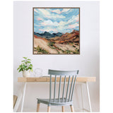 Square framed canvas of a desert landscape with mountains under a blue sky filled with textured white clouds, hanging above a wooden desk and gray chair in a minimalist room.