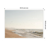 Serene coastal beach scene with gentle waves rolling onto sandy shore under a pale sky dotted with flying birds, framed in white with 38x28 inch dimensions.