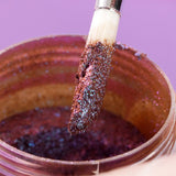 Close-up of a brush coated with purple, glittery pigment powder held above an open jar filled with shimmering violet-colored pigment.