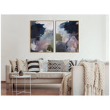 Two abstract framed canvases with navy, purple, and muted green brushstrokes hang above a beige sofa adorned with black and white patterned pillows and a chunky knit throw.