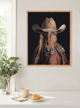 Portrait of a chestnut horse wearing a weathered brown cowboy hat against a black background, framed in light wood and hung on a cream wall.