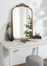 Arched mirror with ornate carved wood top on white vanity holding green glass vases, books, and gold bird sculpture in soft natural light.