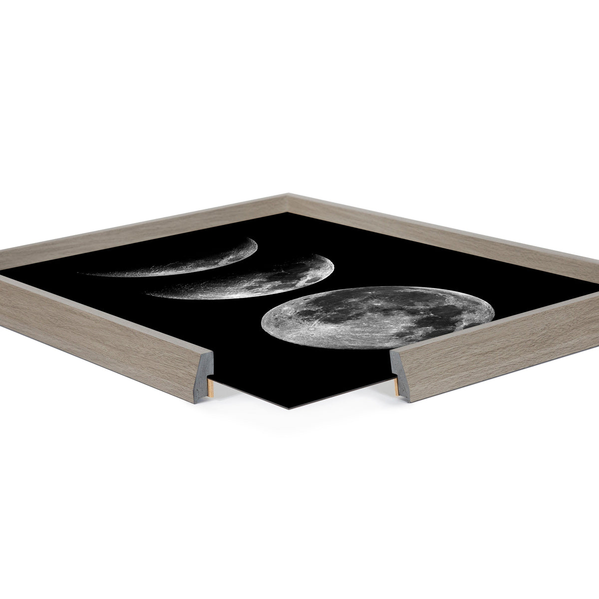 Square framed print depicting four monochrome moon phases from crescent to full against a solid black background in a light wood frame shown at an angle with two corners cut away