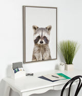 Framed canvas of a young raccoon with detailed fur and dark facial markings, hanging above a white desk with a potted plant and stationery items.