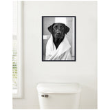 Black and white photograph of a black Labrador dog wrapped in a white bath towel, framed and hanging on a white wall above a white toilet.