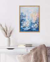 Framed underwater ocean landscape print with blue, coral pink, and soft yellow aquatic plants against a light water surface, hung above a white desk with a vase and books.