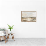 Muted abstract landscape painting with beige, brown, and gray tones showing a cloudy sky and subtle water reflections in a light wood frame on a white wall above a modern side table and sofa.