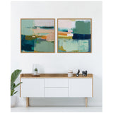 Two framed abstract canvases with layered blocks of green, blue, orange, beige, and pink hues above a white cabinet with wooden top, accentuated by a small plant and decorative items.