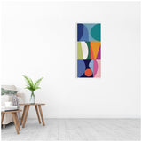 Tall rectangular canvas with colorful geometric abstract shapes including circles and triangles in blue, green, orange, pink, and purple, hanging on a white wall above a wooden side table and beige sofa.