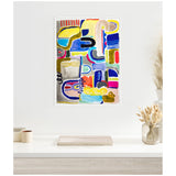 Abstract framed canvas with irregular yellow, blue, pink, and red shapes and lines on a gray and white background, hanging on a white wall above a minimal desk.