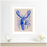 Framed artwork of a blue lobster with detailed claws and legs on a beige background, hanging centered above a white shelf with a glass candle and a small flower vase.