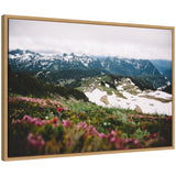 Snow-covered mountain peaks under cloudy sky with green hills and patches of wildflowers in the foreground, viewed through a light wood frame.