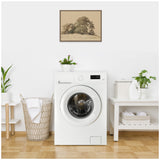 Laundry room with a white front-loading washing machine centered between a wicker laundry basket with a white cloth and a white table holding plants, detergent, and towels below a muted brown framed tree landscape drawing.