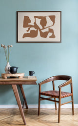 Framed canvas featuring abstract brown distorted shapes on a white background, hanging on a pale blue wall above a mid-century modern wooden chair and table.