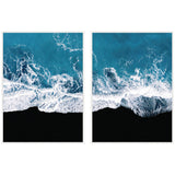 Two framed canvas prints showing aerial views of ocean waves crashing onto a black sandy beach with foamy white water against deep blue sea.