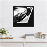 Black and white close-up photo of a vinyl record player spinning a record, framed and hung on a white wall above a minimalist white dresser with plants and books.