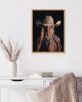 Framed portrait of a brown horse wearing a tan cowboy hat and saddle against a dark background, hanging above a white desk with books and a vase with pampas grass.