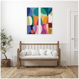 Two-piece abstract canvas art featuring geometric shapes in blue, green, orange, pink, and white, displayed above a wooden framed white cushion bench in a minimalist room.