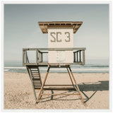 White lifeguard tower labeled SC 3 with gray railings and wooden stairs standing on light sandy beach under pale blue sky and calm ocean horizon