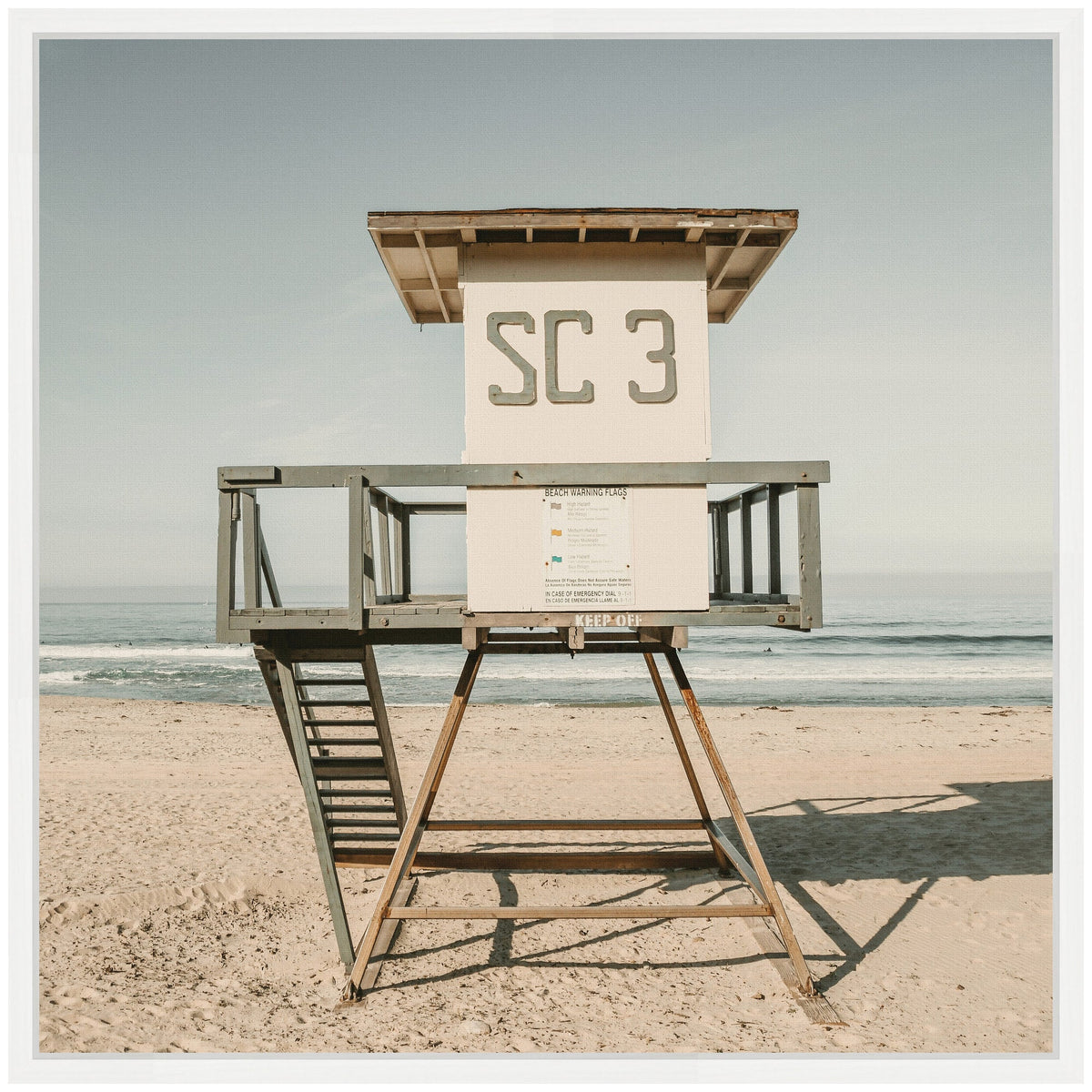 White lifeguard tower labeled SC 3 with gray railings and wooden stairs standing on light sandy beach under pale blue sky and calm ocean horizon