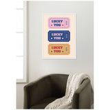 Framed wall art featuring three rectangular tickets in pink, blue, and mustard with 'LUCKY YOU' and star accents stacked vertically against a white wall above an olive green chair with a knitted throw.