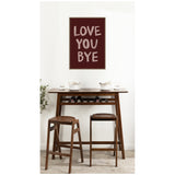 Framed maroon canvas with pink hand-painted text 'LOVE YOU BYE' hangs above a tall dark wood table with two matching stools and white dishes in a simple room.