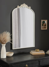 Arched white-framed wall mirror with ornate scrollwork at the top, hanging above a black console with neutral decor and a small framed photo on a dark wall.