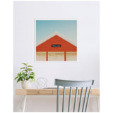 Framed square canvas print of a red and white peaked roof against a clear blue sky, hanging on a white wall above a light wood desk with a potted plant and chair.